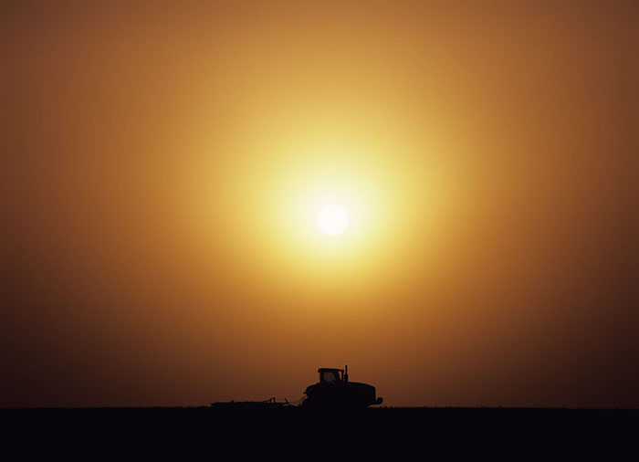 A caterpillar pulling discs sits idle and in silhouette at the crest of a hill as the sun moves toward the horizon. &nbsp;This...